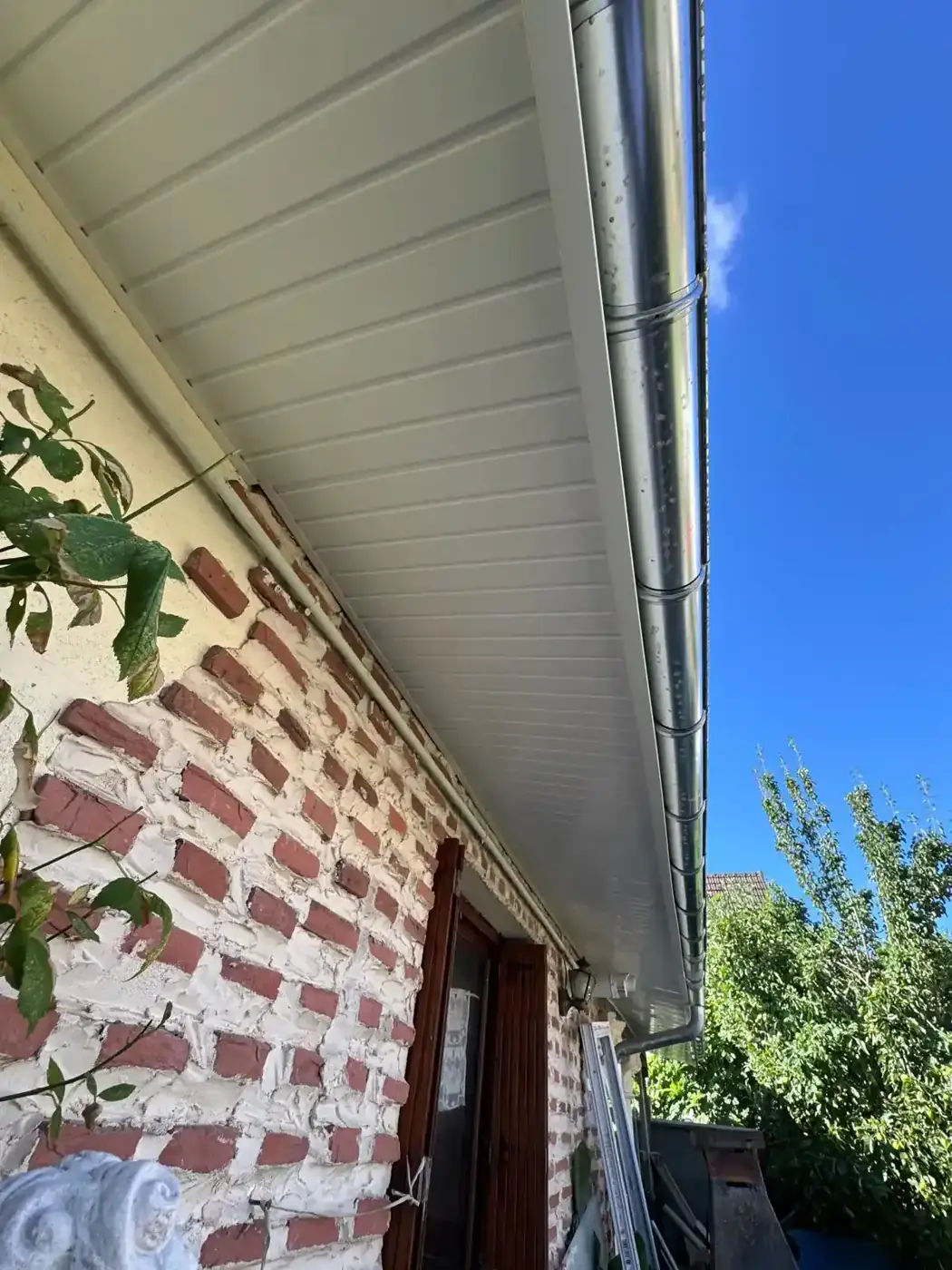 Sous-face de débord de toiture en lames PVC blanc emboîtées et gouttière demi-ronde en zinc posées sur un mur en briques à Chelles